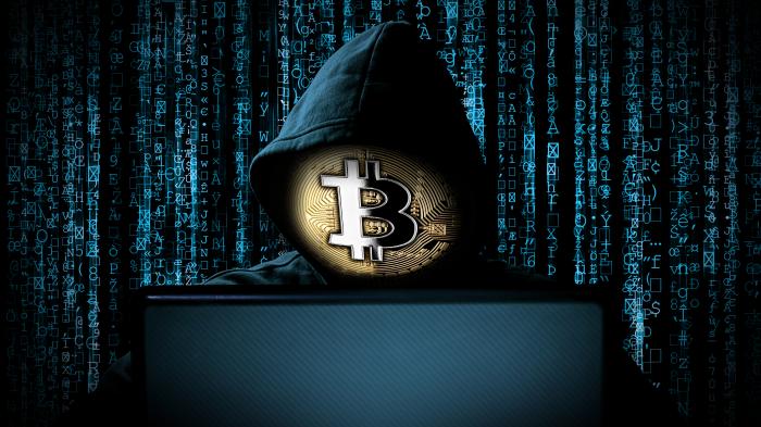 Person in Kaputzenpulli sitzt im dunklen vor einem Laptop; statt dem Gesicht der Person ist eine güldene Münze mit Bitcoin-Logo zu sehen.